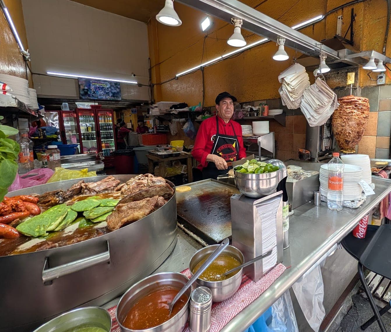 A taquero working the grill at a Mexico City taquería, with a trompo of al pastor, cazuelas of stewed meats, and fresh salsas
